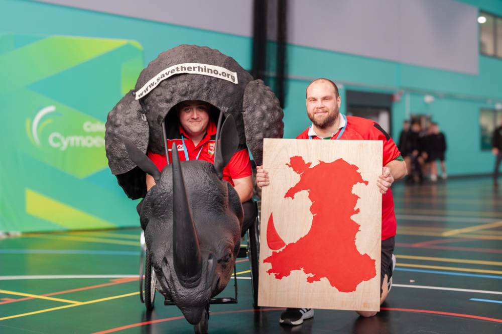 Two men in Wales rugby league shirts, one in the rhino costume chair, another by his side, with a red map of Wales.