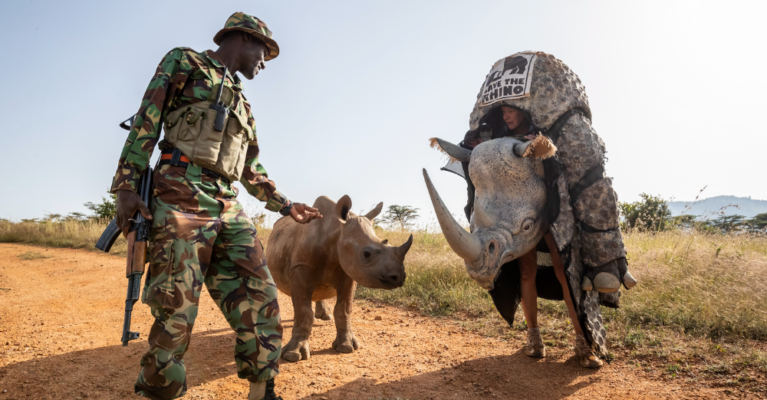 A woman in a rhino costume, next to a black rhino calf and a ranger, outside
