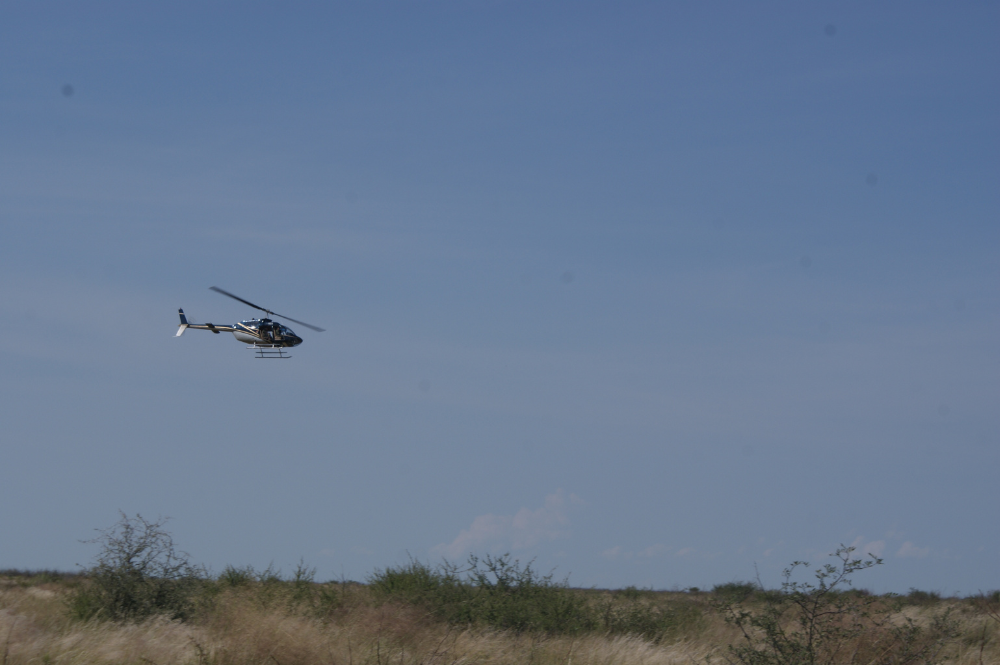 A helicopter in the sky flying over a green landscape.