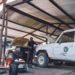 A mechanic fixing two cars in a workshop outside.