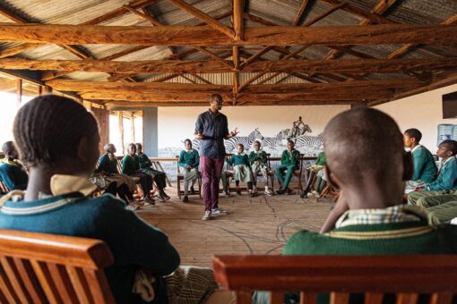 Man standing smiling leading a group of people sitting in a circle around him.