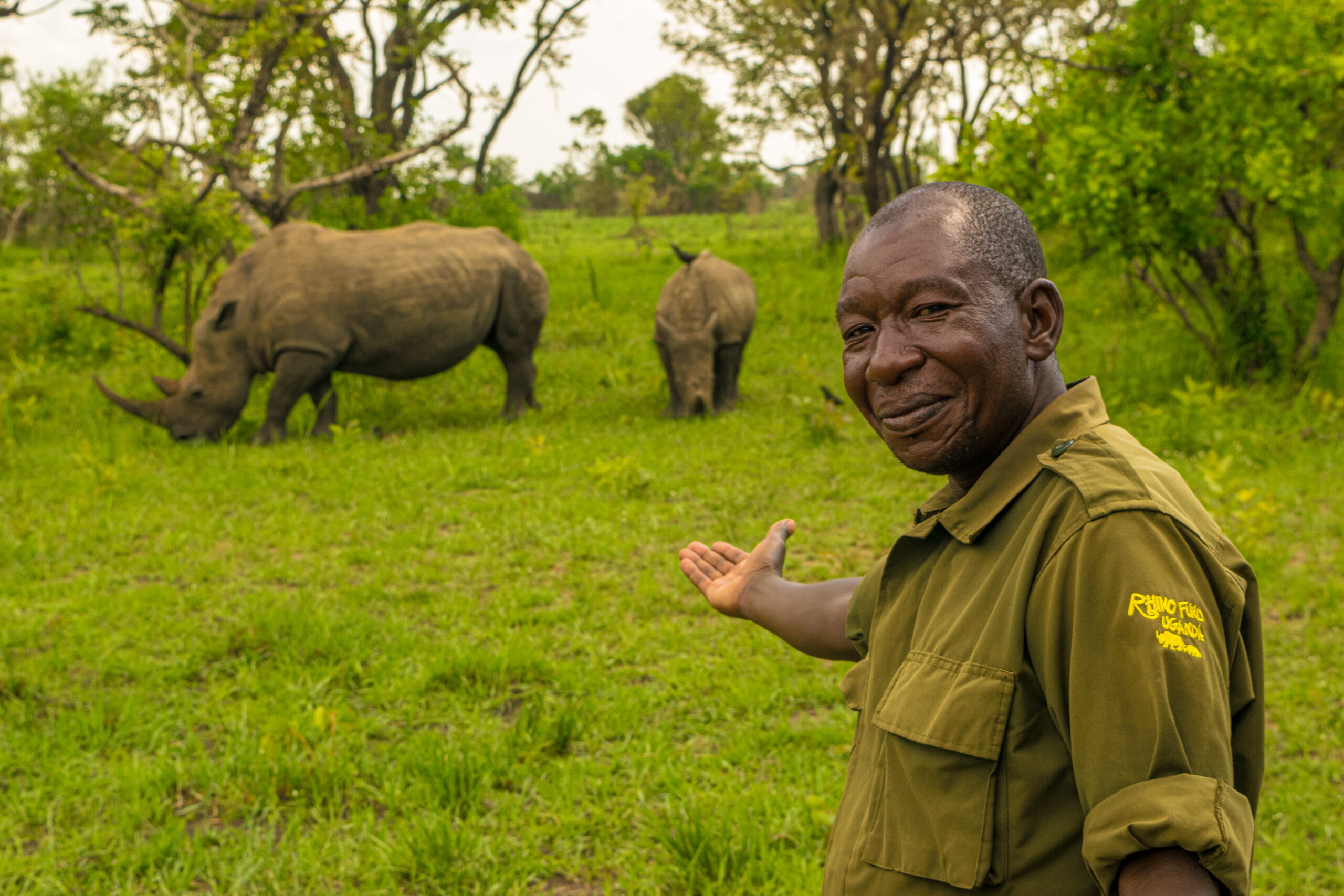 A ranger smiles at the camera while pointing to 2 rhinos happily grazing behind him